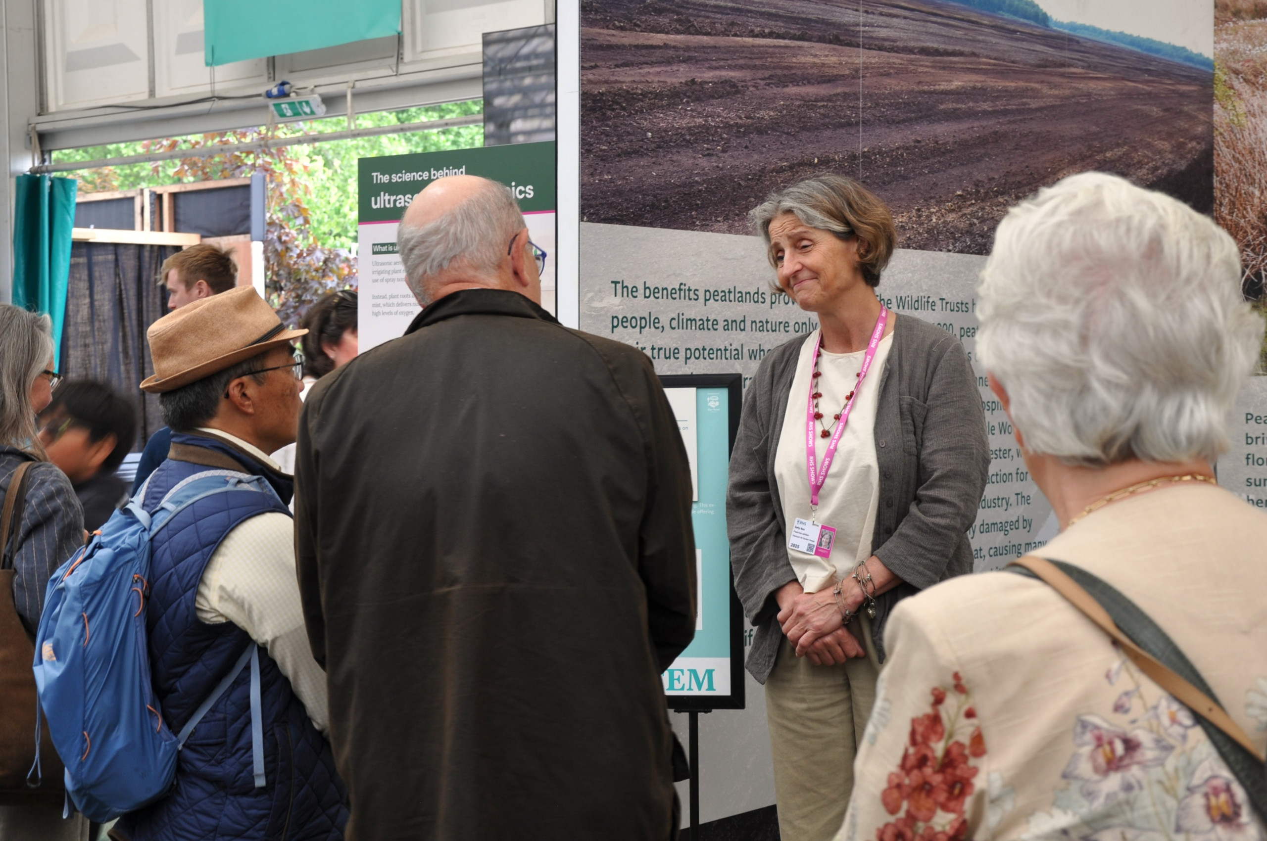 Peat-free Partnership Advocate Sally Nex discusses peat-free growing with visitors at Chelsea Flower Show. Sally is a middle-aged white woman with tan skin and dark grey hair.