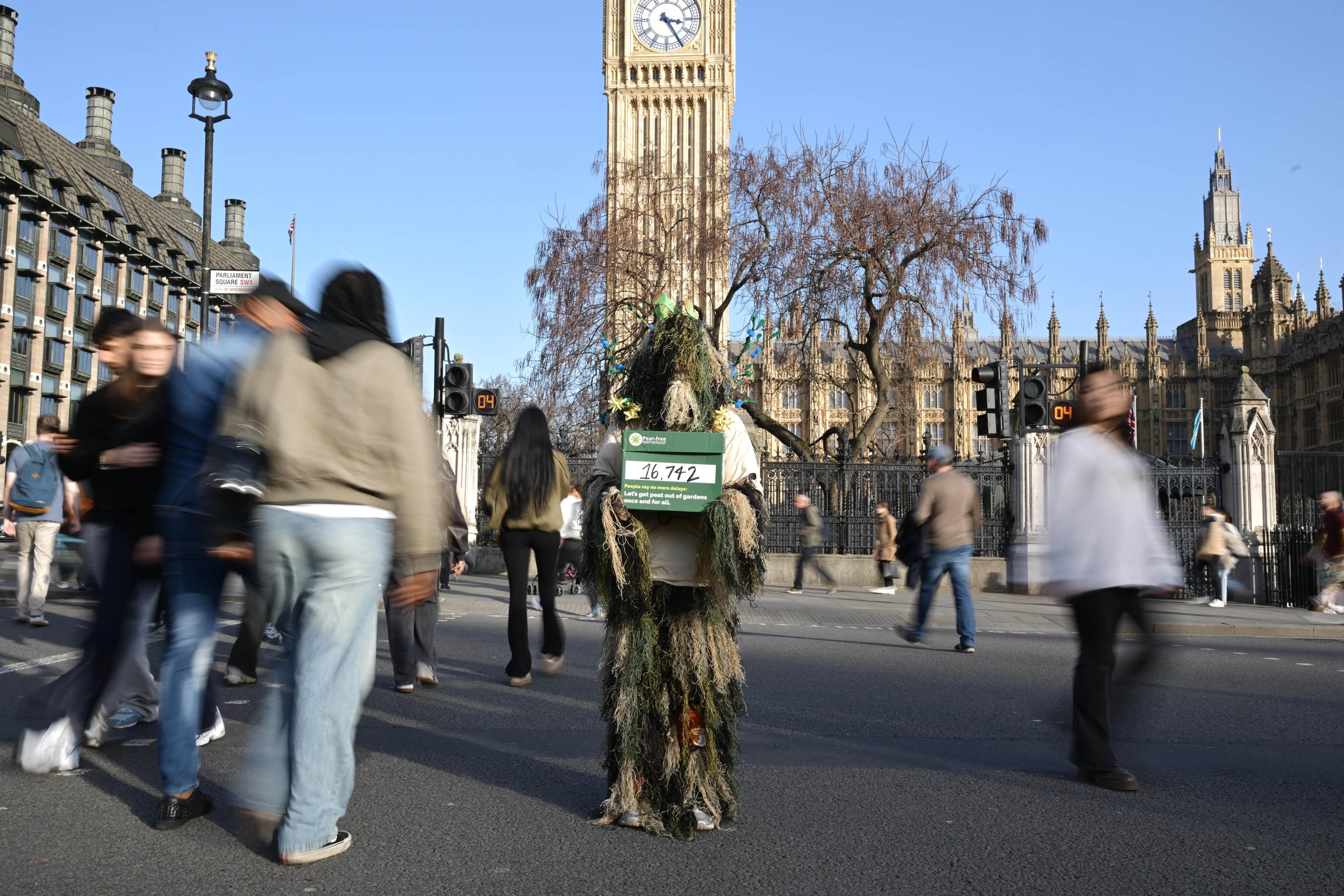 A person in a bog creature costume stands in a crossing in front of Big Ben. Blurry passersby continue with their lives, ignoring the bog creature.