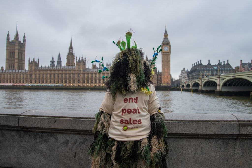 Photograph shows a 'bog creature' covered in moss and peatland plants, standing with the River Thames behind them and the Houses of Parliament in the background. The creature wears a t-shirt that reads 'end peat sales'