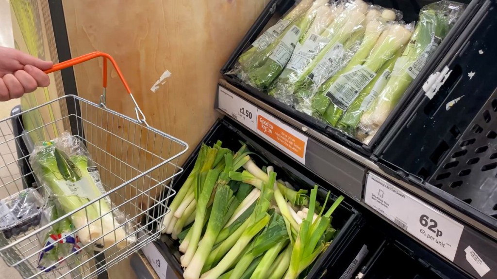Photograph shows a persons hand holding a shopping basket with orange handles. They are in the fresh fruit and vegetables aisle and have both blueberries and leeks in the basket.